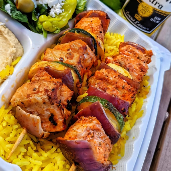 Chicken and vegetable kebabs on yellow rice with hummus and salad in a takeout container.